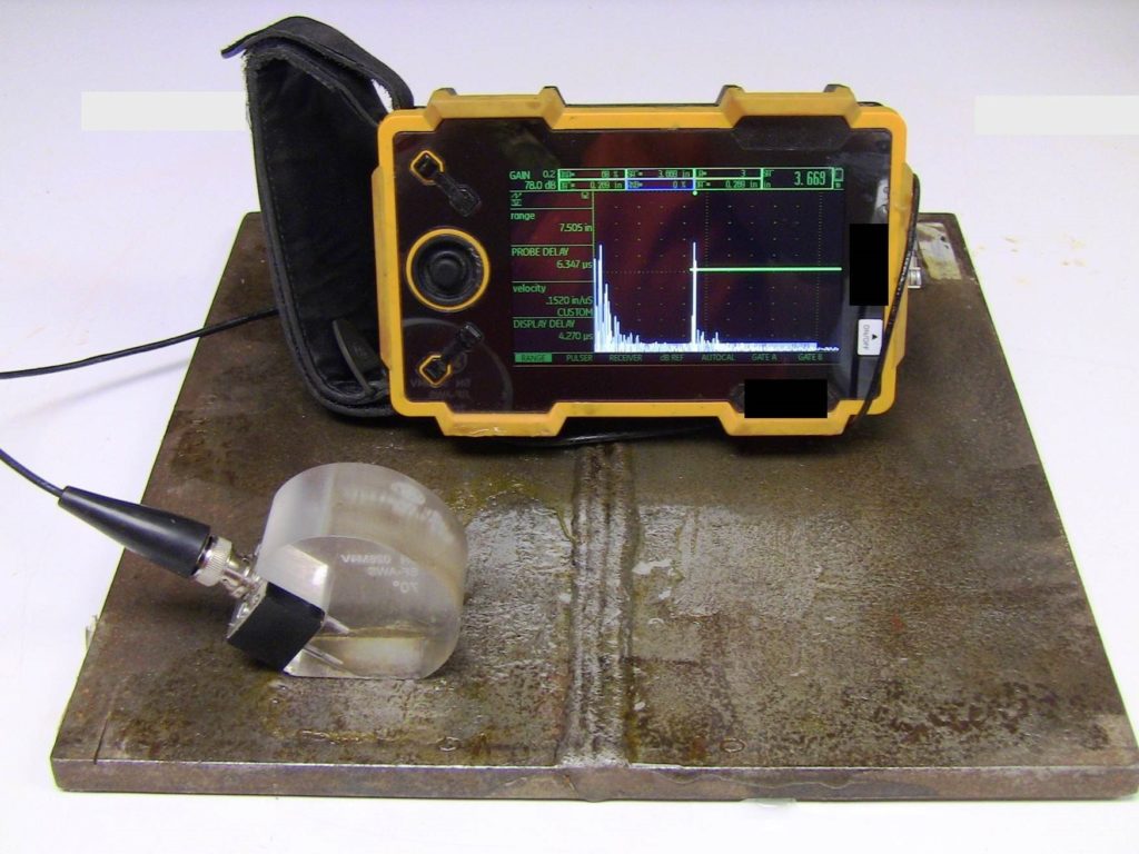 An angled-beam transducer and its control panel are on the surface of a rectangular test plate. The transducer is resting on the lower left corner of the test plate. The control panel is resting on the approximate center of the plate.