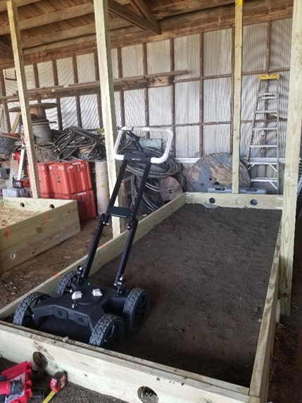 A GPR system in a four-wheeled cart with a handle sits inside a wood-framed box filled with soil up to the drilled holes.