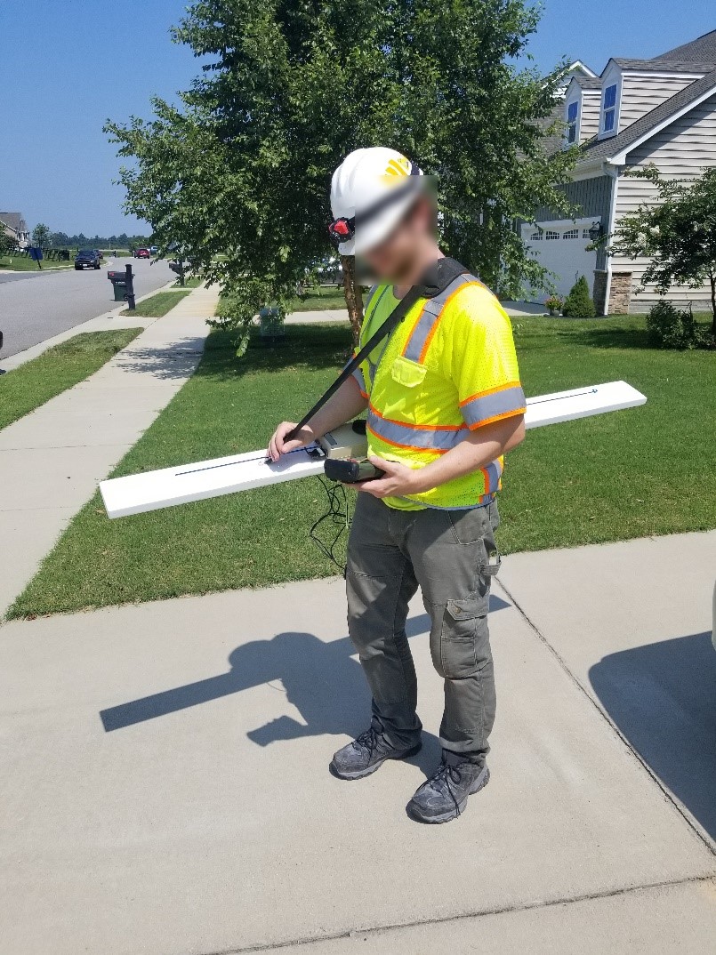 An operator is standing in the driveway of a home in a subdivision holding an FDEM conductivity meter. A multifrequency EM sensor boom is attached to a strap over his shoulder and is hanging parallel to the ground.