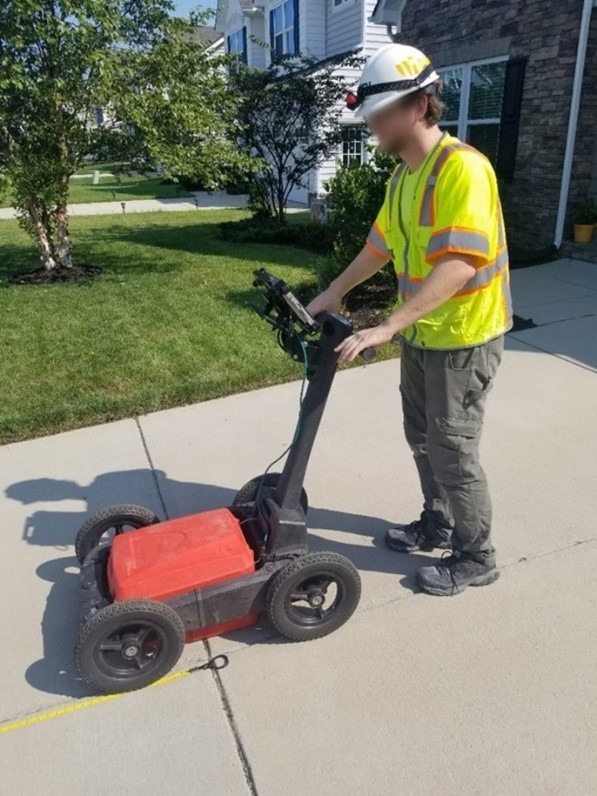 A worker pushes a GPR system, which is situated in the center of a four-wheeled cart with a handle, on a concrete driveway.